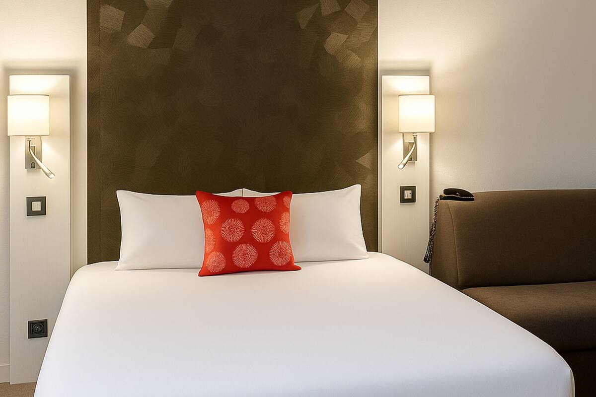 Una habitación de hotel con una cama grande, dos lámparas de noche, un cojín rojo estampado y un sofá marrón.