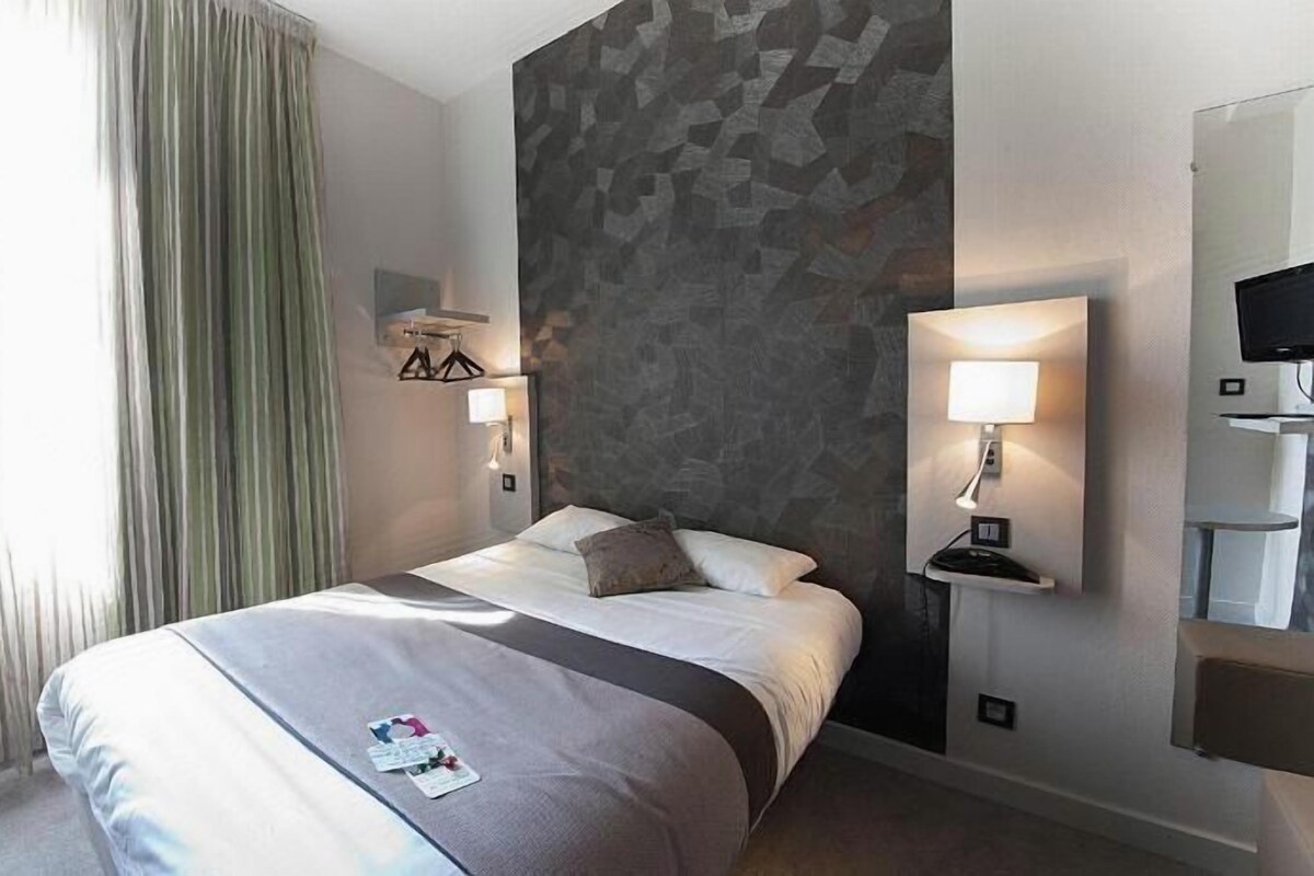 Une chambre d’hôtel avec un lit, deux tables de chevet, un téléviseur fixé au mur et une fenêtre avec des rideaux.