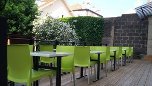 Une rangée de tables et de chaises d’extérieur, avec une terrasse en bois, une haie verdoyante et un mur de briques en arrière-plan.