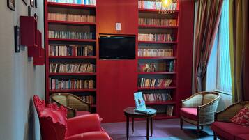 A cozy living room with a red sofa, a wooden armchair, and a bookshelf filled with books. A flat-screen TV is mounted on the wall, and a chandelier hangs from the ceiling.