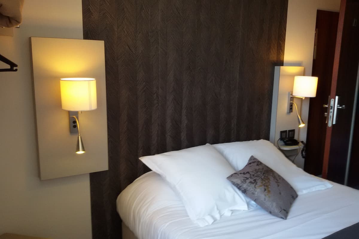 Una habitación de hotel con una cama, dos lámparas fijadas a la pared, un cabecero de madera y una puerta.
