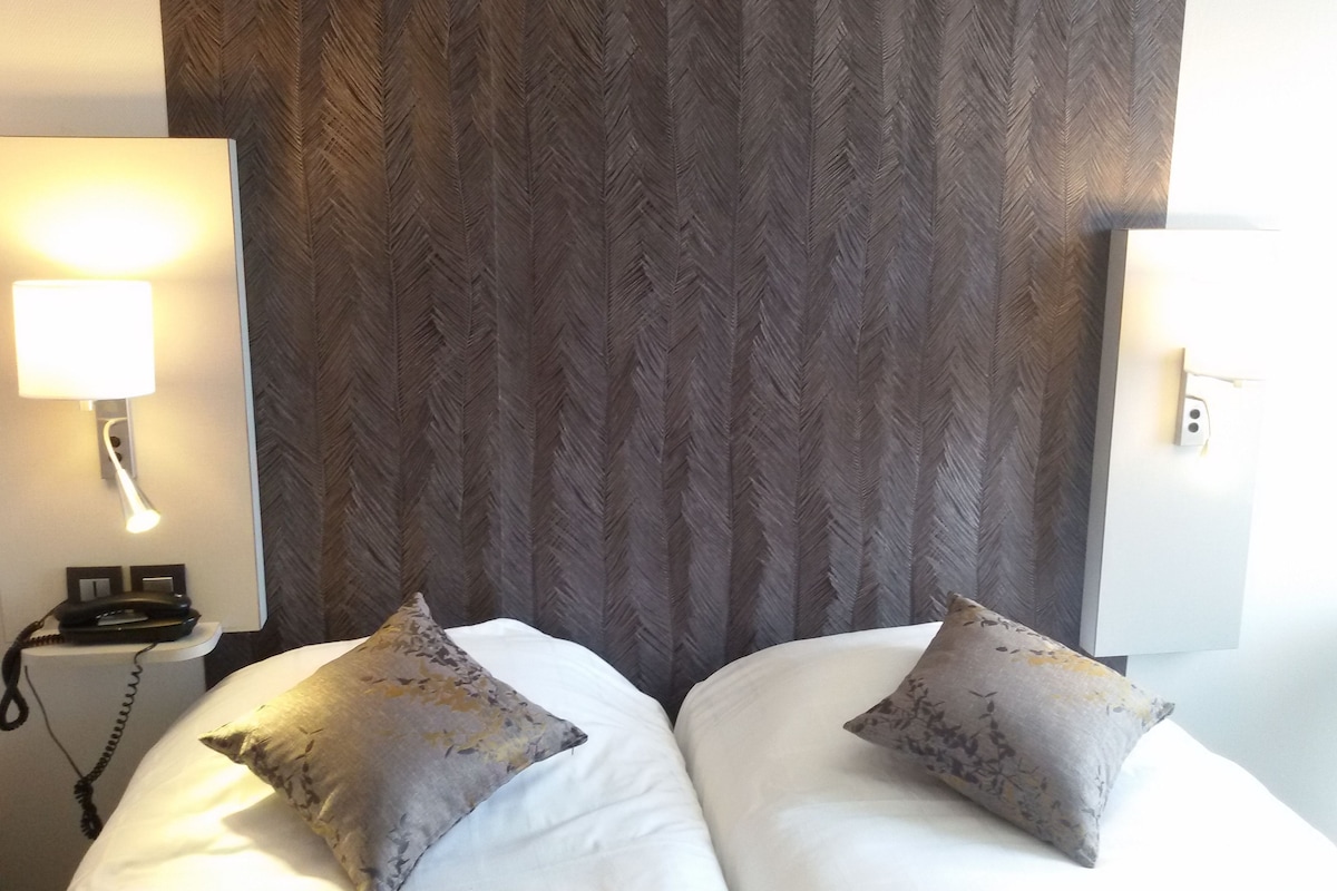 Una habitación de hotel con dos camas, una mesita de noche con lámpara y un papel tapiz estampado.