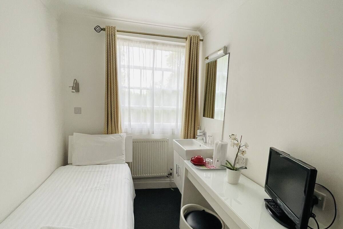 Una habitación de hotel con cama, escritorio con ordenador, radiador y ventana con cortinas.