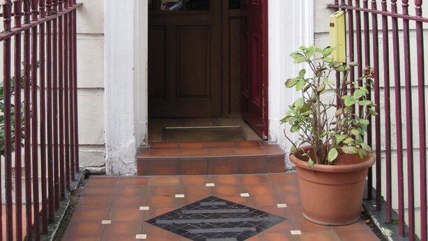 A hotel entrance with a red door, a potted plant, and a sign indicating a warning for a steep staircase. - Meridiana (London)