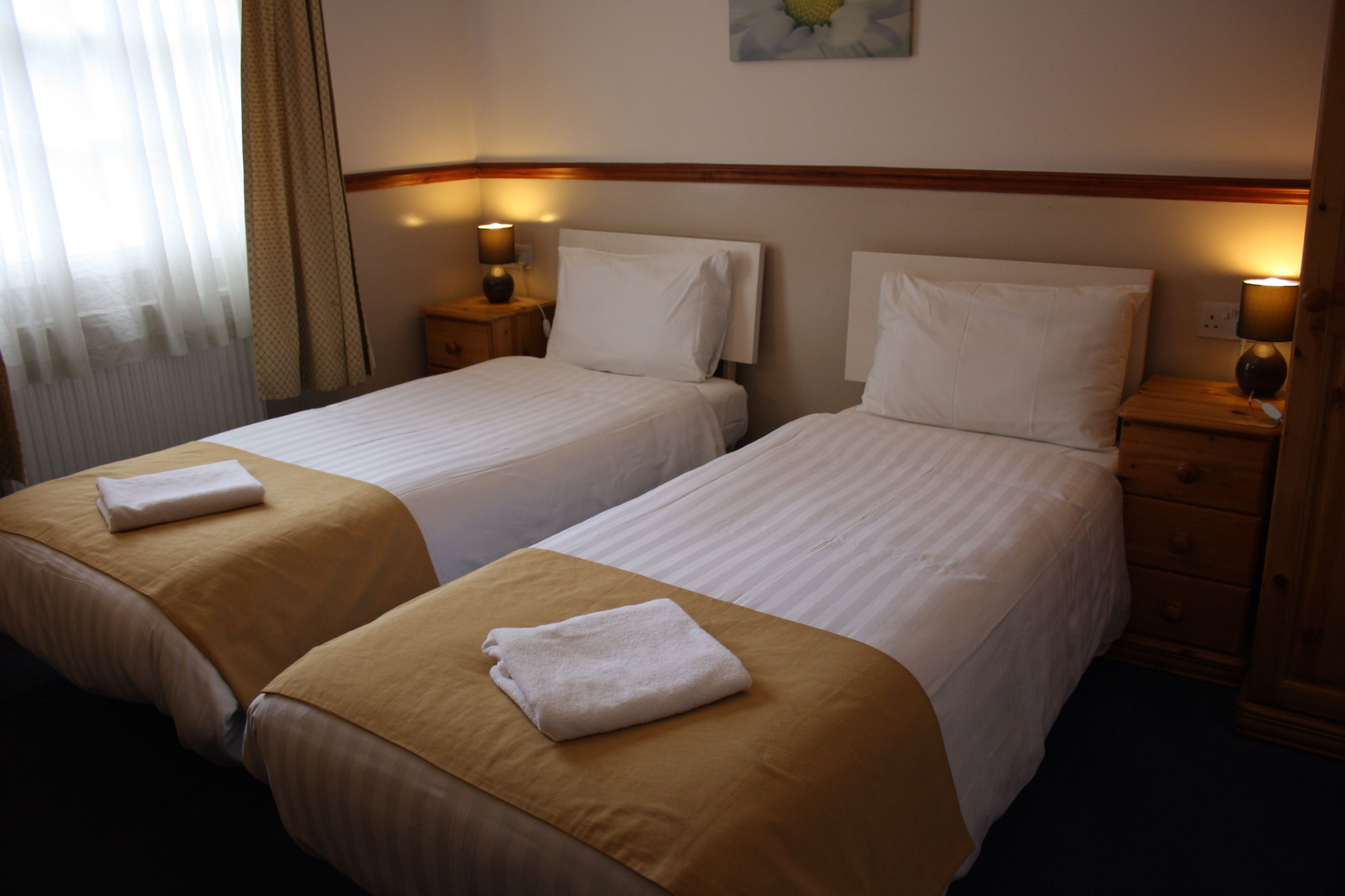 Una habitación de hotel con dos camas individuales, cada una con un edredón blanco y una toalla blanca doblada. Hay mesitas de noche de madera con lámparas a cada lado de las camas.
