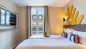 Une chambre d’hôtel avec un grand lit, un ordinateur portable posé sur le lit, un téléviseur accroché au mur et une vue sur la ville par la fenêtre.