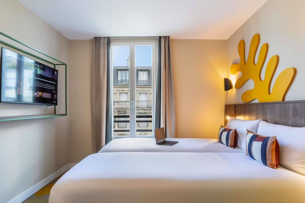 A hotel room with a large bed, a laptop on the bed, a TV on the wall, and a view of the city through the window.