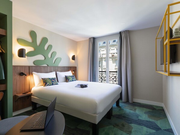 A hotel room with a bed, a laptop on a table, a wall-mounted light, and a view of buildings through the window. - ibis Styles Paris Jardin de la Villette (Paris)