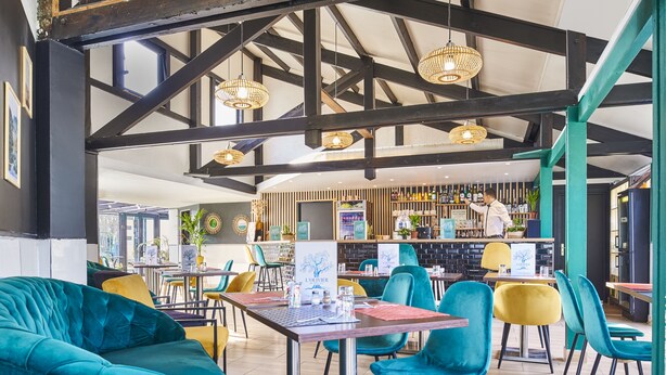 A restaurant with a high ceiling, exposed beams, and hanging wicker lamps. There are tables and chairs with a mix of colors, and a bar area in the background. - ibis budget Saint Ouen l'Aumône (Saint-Ouen-l'Aumone)