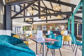 Un restaurant avec un plafond haut, des poutres apparentes et des lampes en osier suspendues. On y trouve des tables et des chaises aux couleurs variées, ainsi qu’un bar situé au fond de la salle.