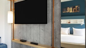 A modern hotel room with a large flat-screen TV mounted on a wooden and metal frame, a bedside table with a lamp, and a bed with a blue headboard. - ibis budget Saint Ouen l'Aumône (Saint-Ouen-l'Aumone)