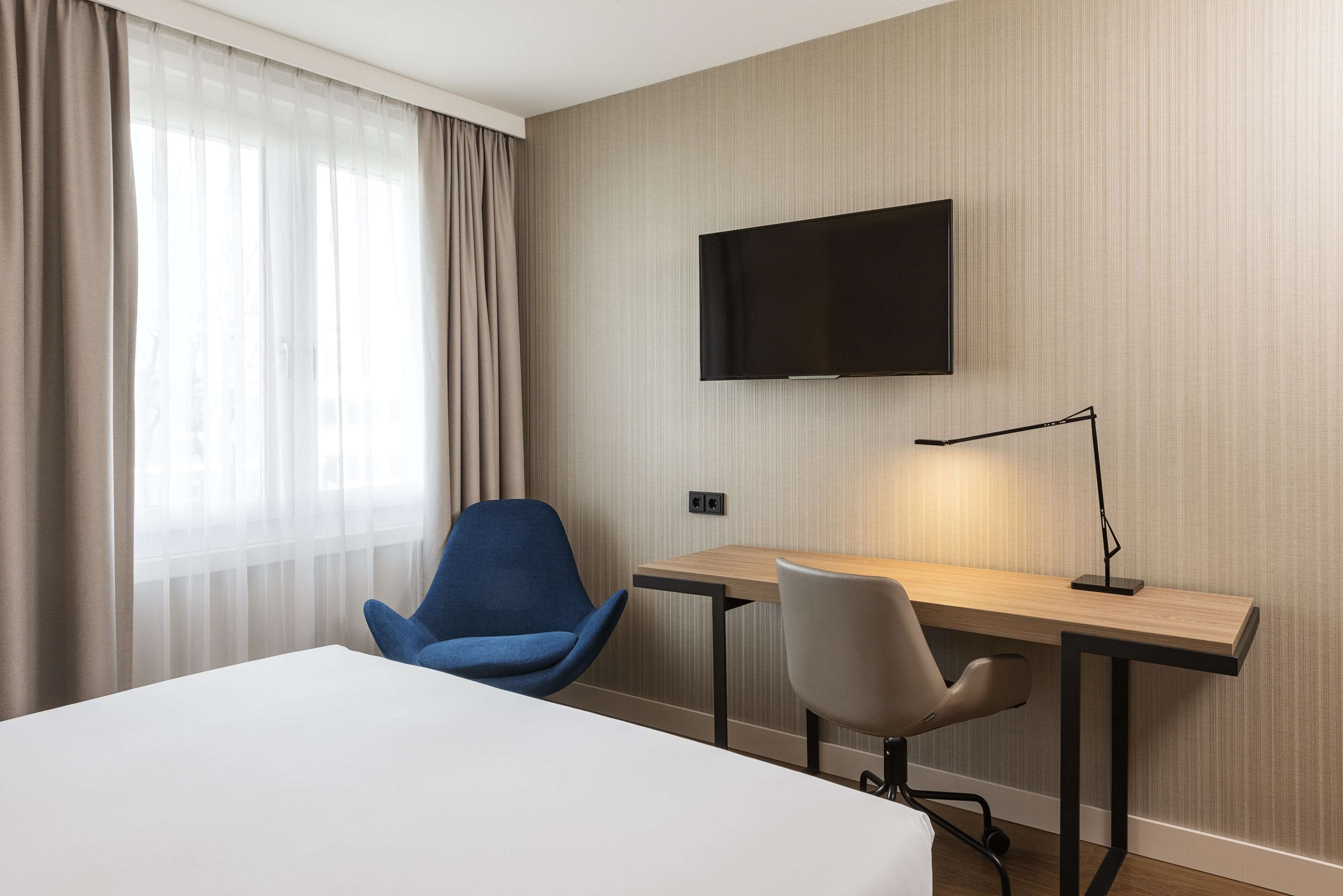 Una camera d'albergo con un letto, una scrivania, due sedie, una TV a muro e una finestra con tende.