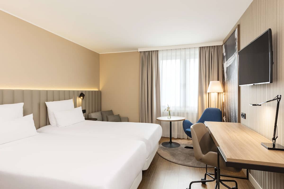 A hotel room with a large bed, a desk, a chair, a TV, and a small table with a lamp.