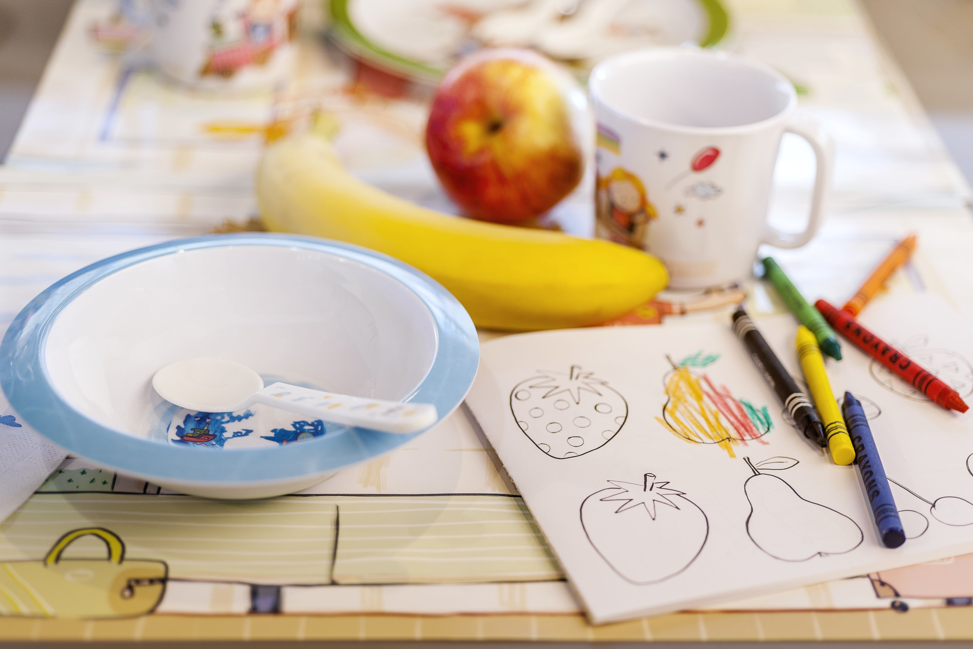 Una mesa con un tazón, cuchara, plátano, manzana y una taza con motivos frutales.