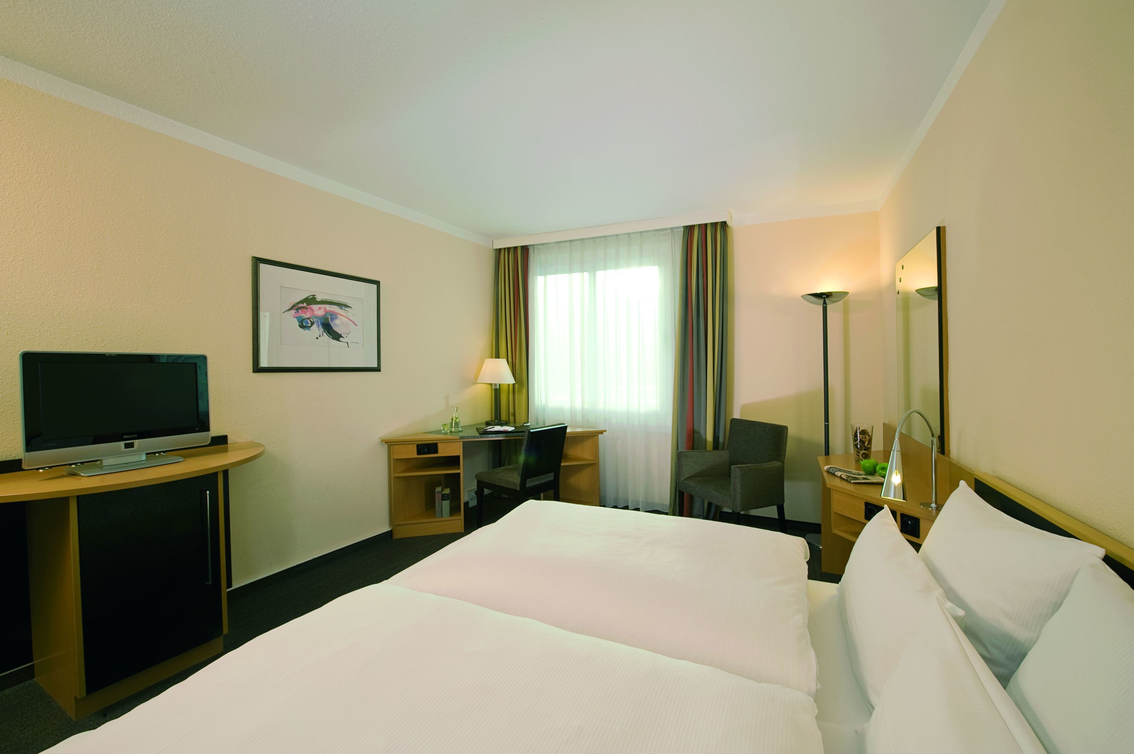 Una habitación de hotel con cama, escritorio, silla, televisor, lámpara y espejo.
