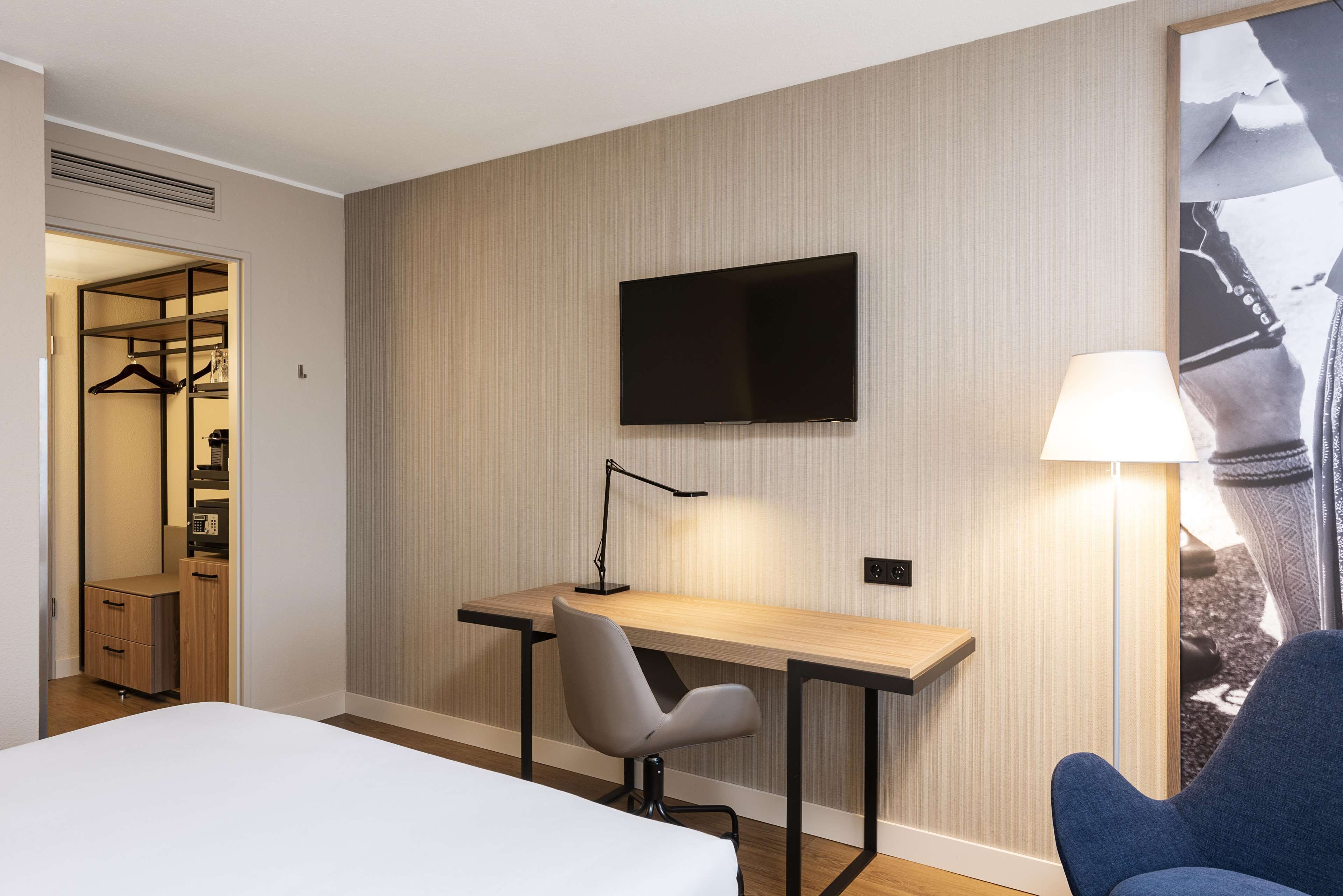 A hotel room with a bed, a desk with a chair, a TV mounted on the wall, and a lamp.