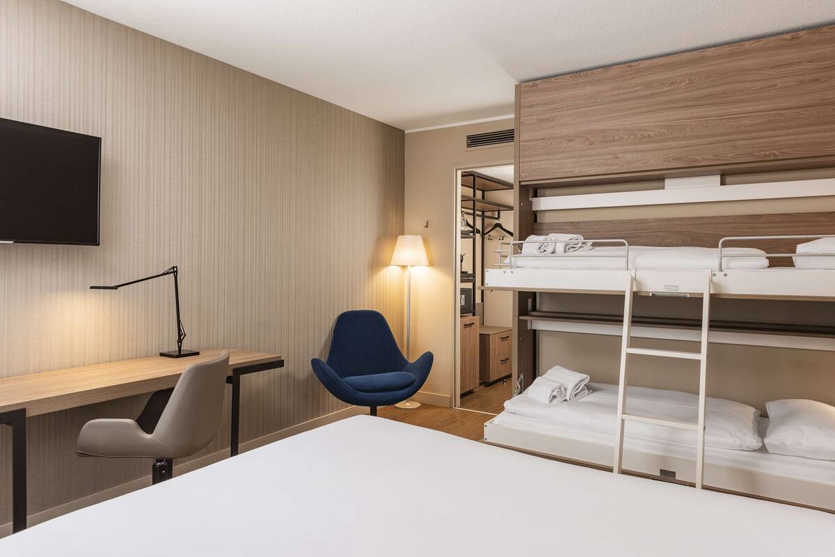 A hotel room with a bunk bed, a desk with a chair, a TV mounted on the wall, and a lamp.