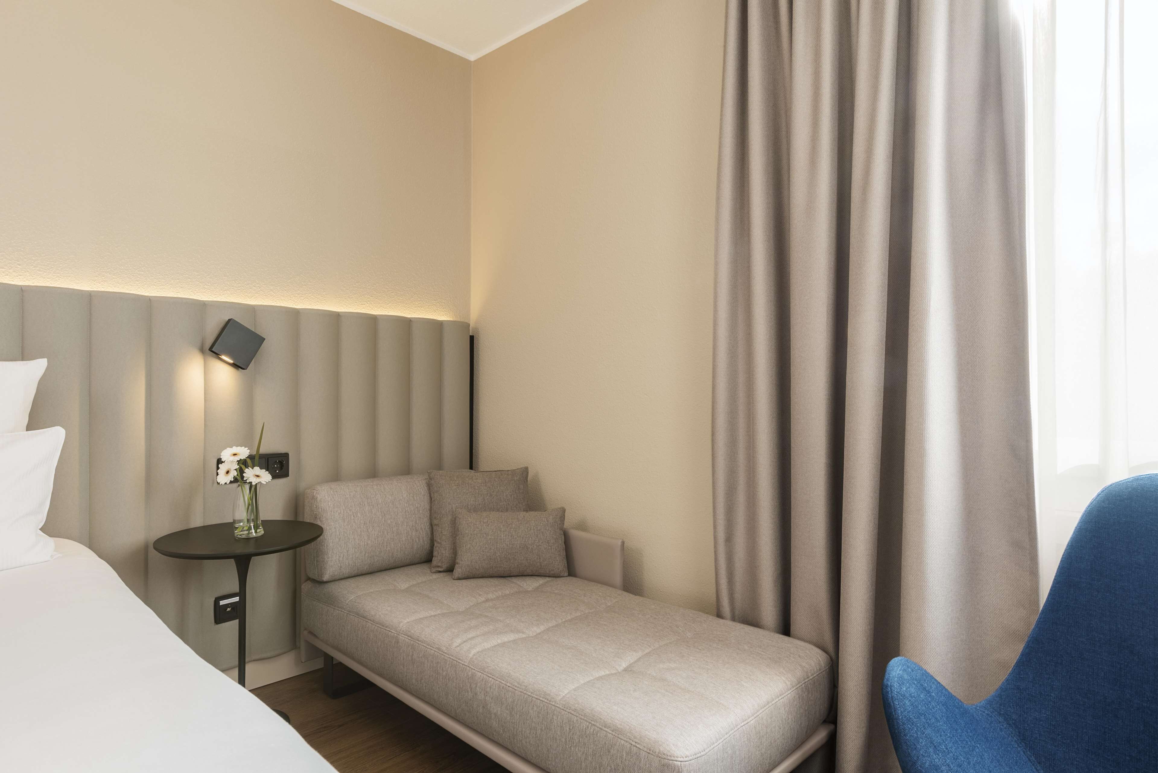 Habitación de hotel con cama, un pequeño sofá, una mesita de noche con un florero y una silla azul.