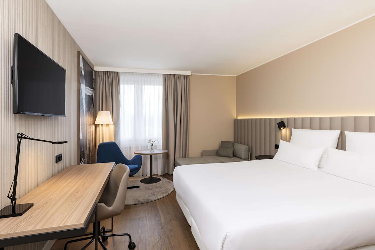 A hotel room with a large bed, a desk with a chair, a TV mounted on the wall, and a small table with a lamp.
