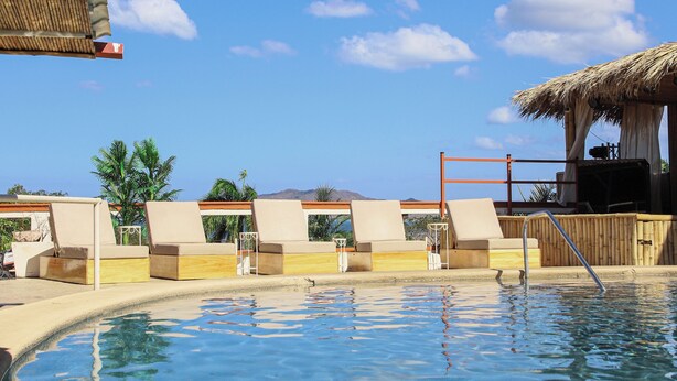 Cielo azul despejado, piscina con sillas de descanso y una estructura de techo de paja.