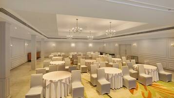 A large banquet hall with round tables and white chairs, chandeliers, and a carpeted floor.