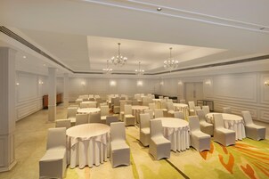 A large banquet hall with round tables and white chairs, chandeliers, and a carpeted floor.
