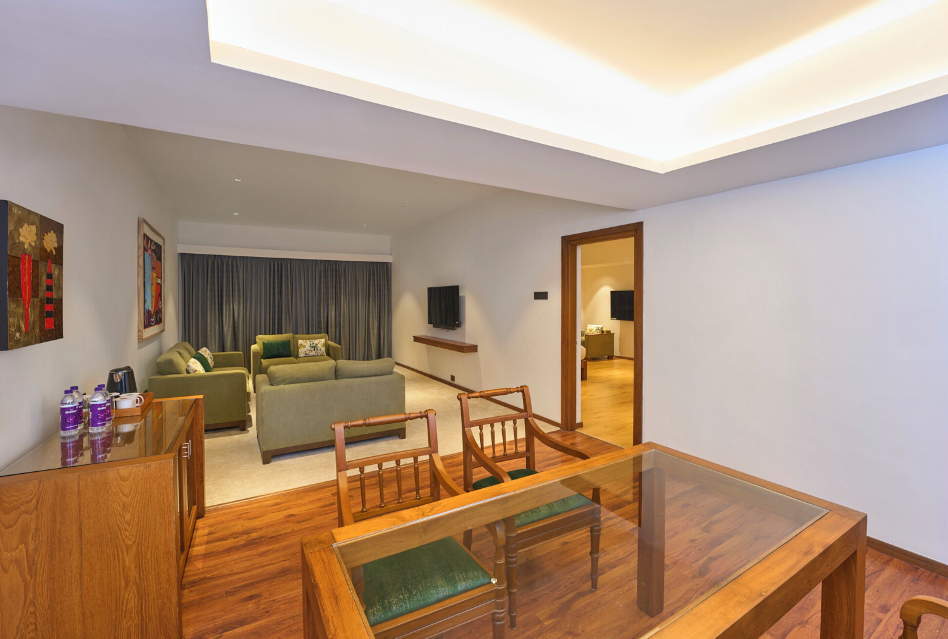 A modern living room with a wooden dining table, green upholstered sofa, and a flat-screen TV mounted on the wall.