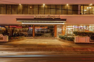 Exterior del hotel Avenue Regent, con una entrada destacada, fachada de vidrio y señalización visible de comercios cercanos.