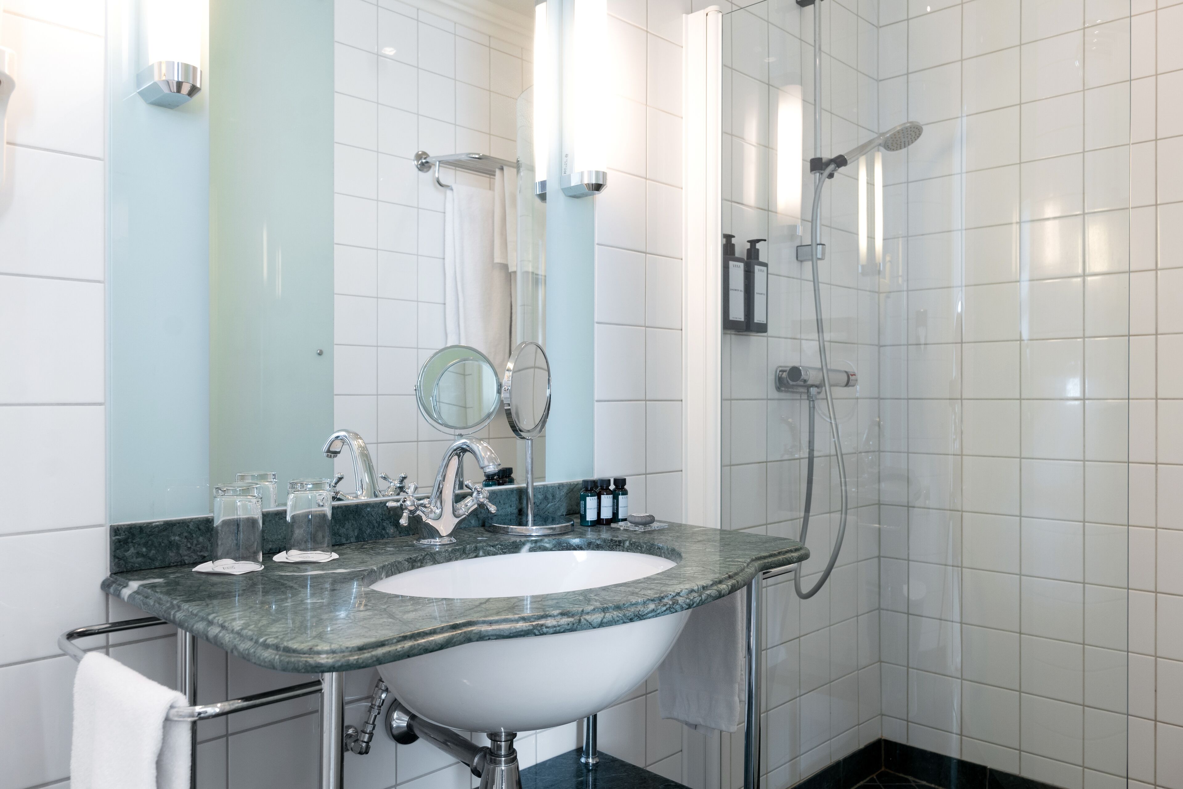 A modern bathroom with a marble sink, two mirrors, a handheld showerhead, and a glass-enclosed shower.