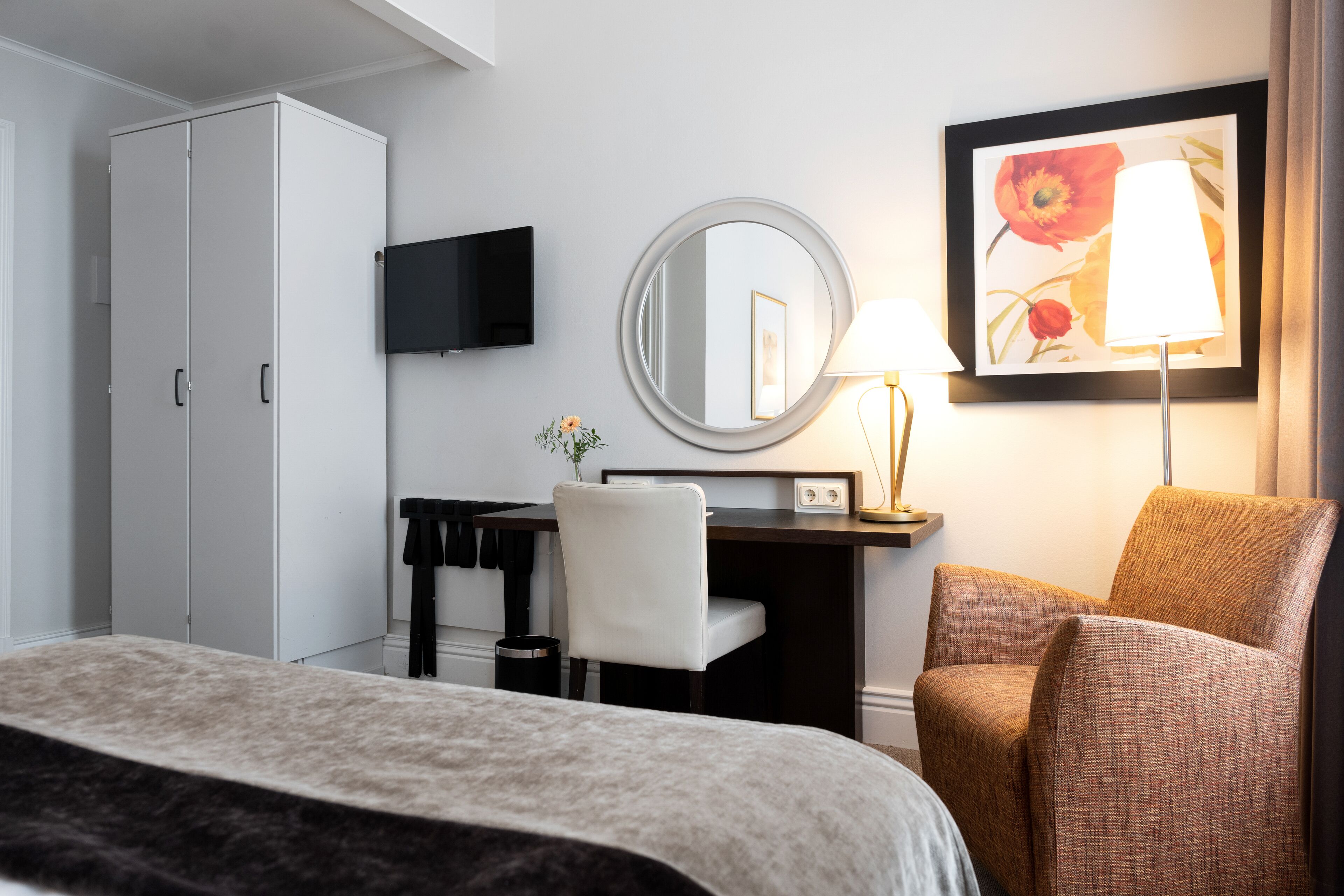 Una habitación de hotel con cama, escritorio, silla, armario, televisor, espejo, lámpara y un cuadro en la pared.