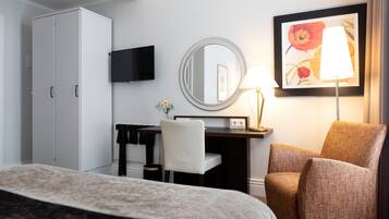 Una habitación de hotel con cama, escritorio, silla, armario, televisor, espejo, lámpara y un cuadro en la pared.
