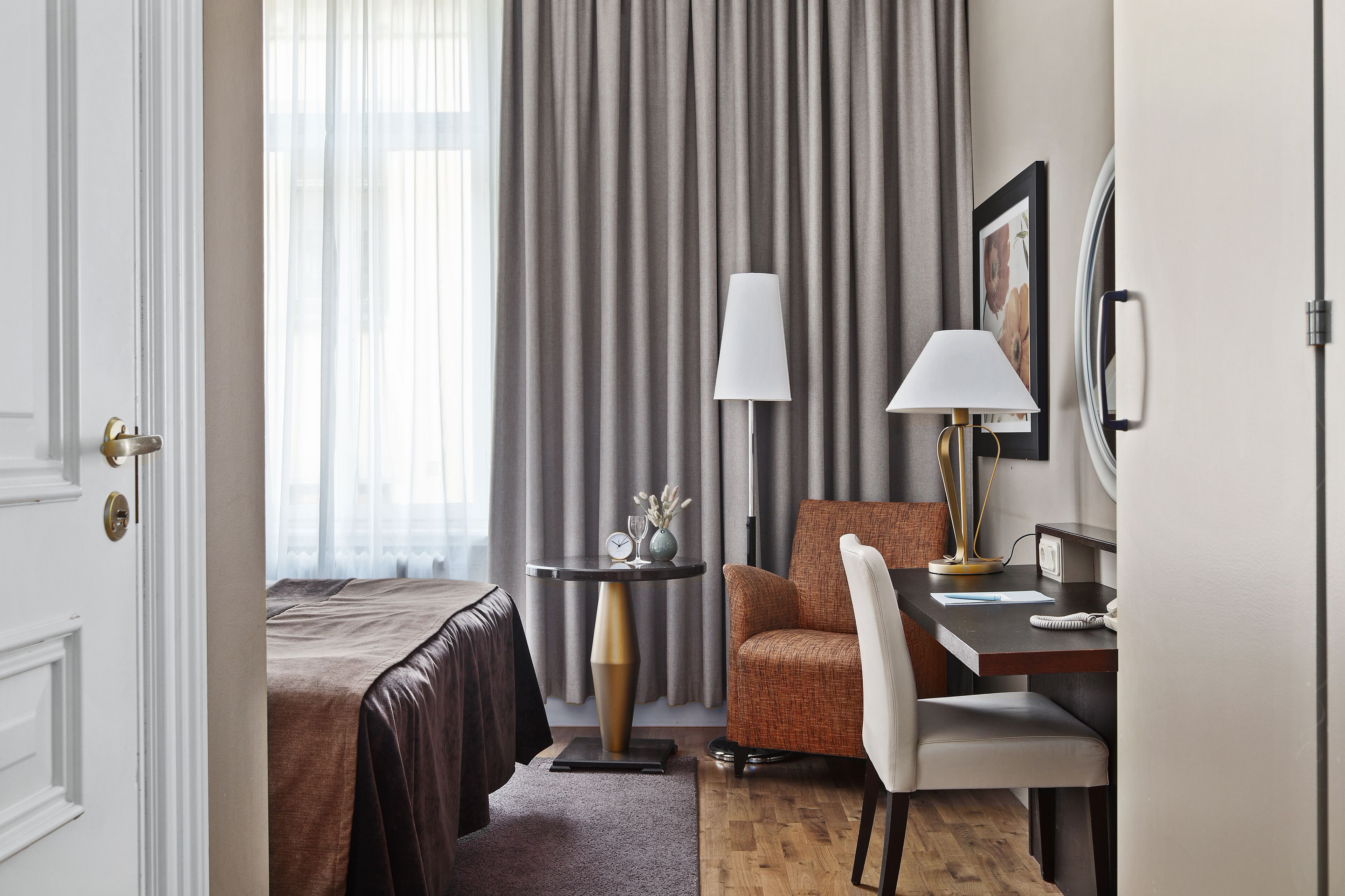 A hotel room with a bed, a desk, a chair, a lamp, and a small table with a clock and decorative items.