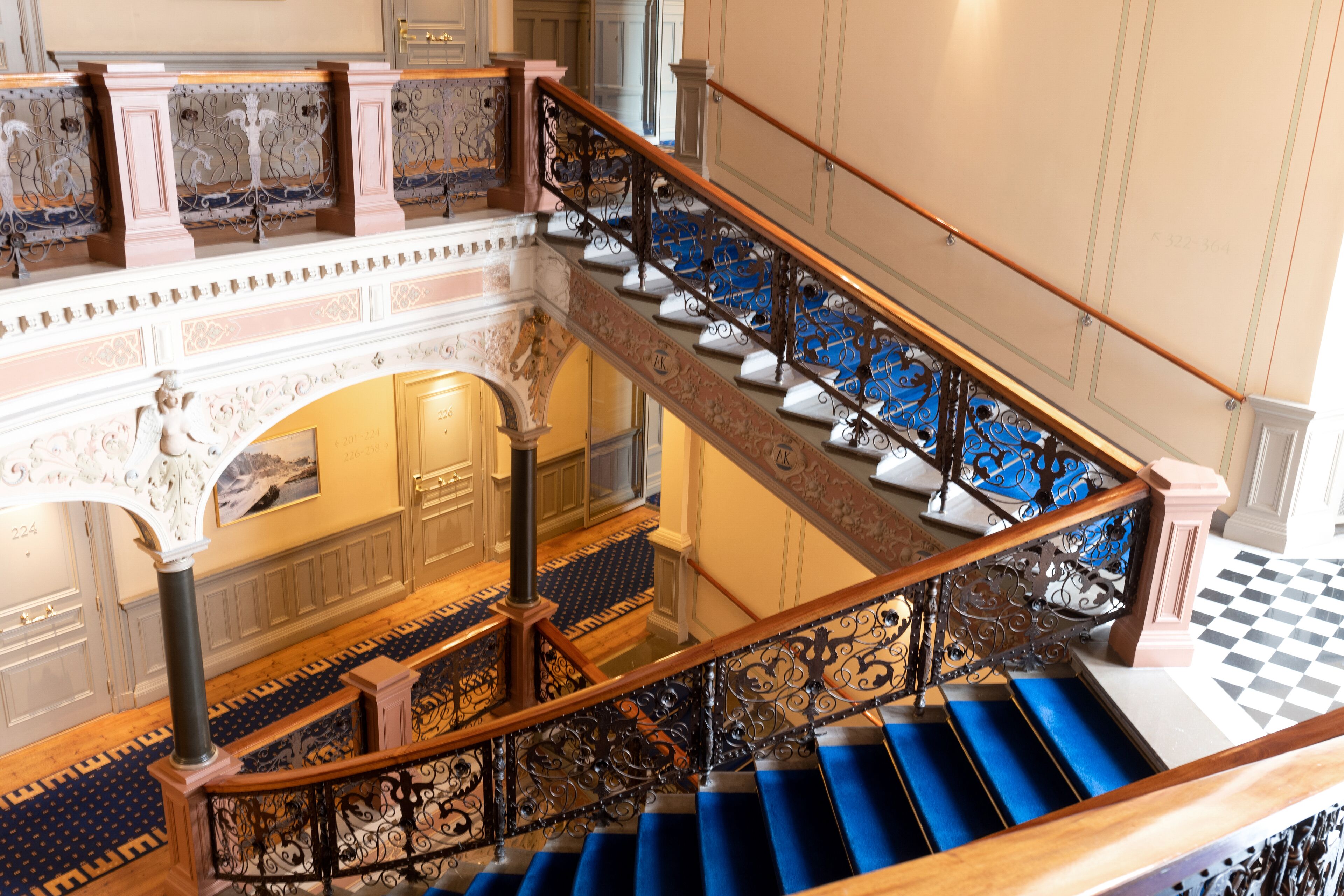 A grand staircase with blue steps and ornate metal railings, leading to an upper floor with a checkered floor and a view of a room with a large window.