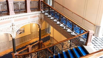 Una imponente escalera con peldaños azules y barandillas de metal ornamentadas, que conduce a un piso superior con suelo de cuadros y vista de una habitación con un amplio ventanal.