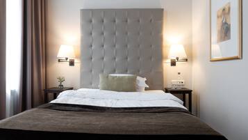 Habitación de hotel con un gran cabecero capitonado, dos lámparas de noche, una mesita de noche con un florero y un cuadro enmarcado en la pared.