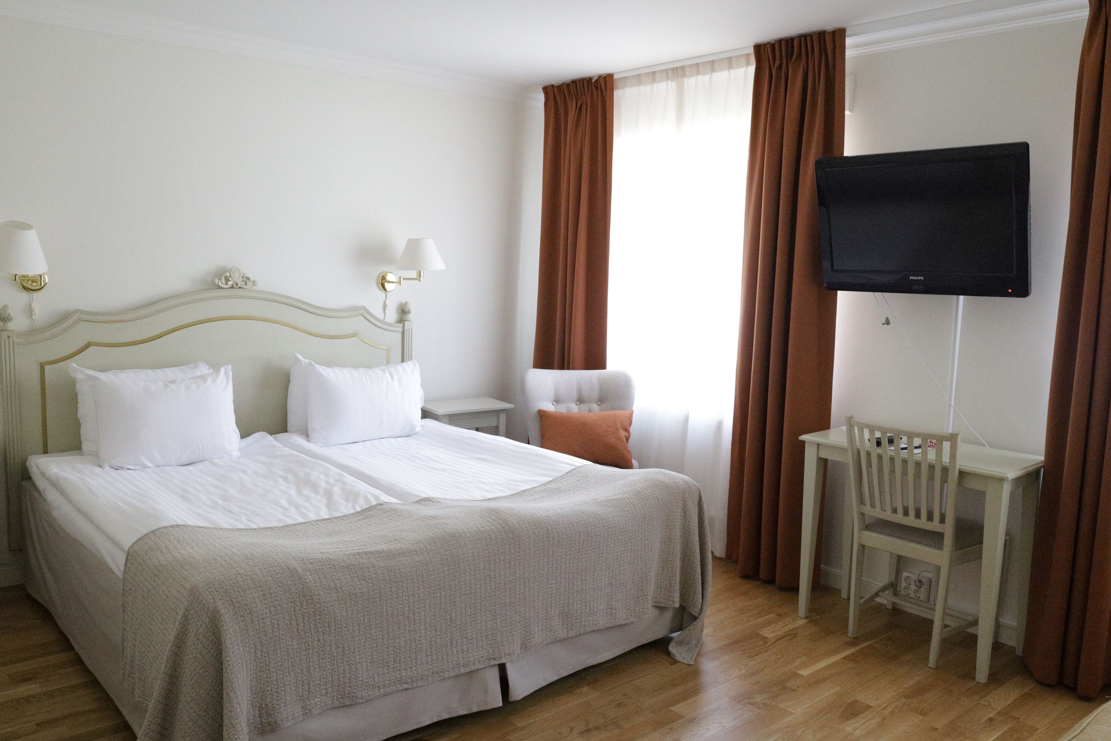 Habitación de hotel con una cama grande, dos mesitas de noche con lámparas, una silla, un televisor montado en la pared y un ventanal con cortinas.