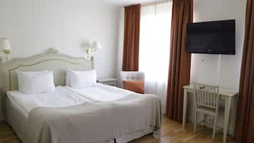 Habitación de hotel con una cama grande, dos mesitas de noche con lámparas, una silla, un televisor montado en la pared y un ventanal con cortinas.