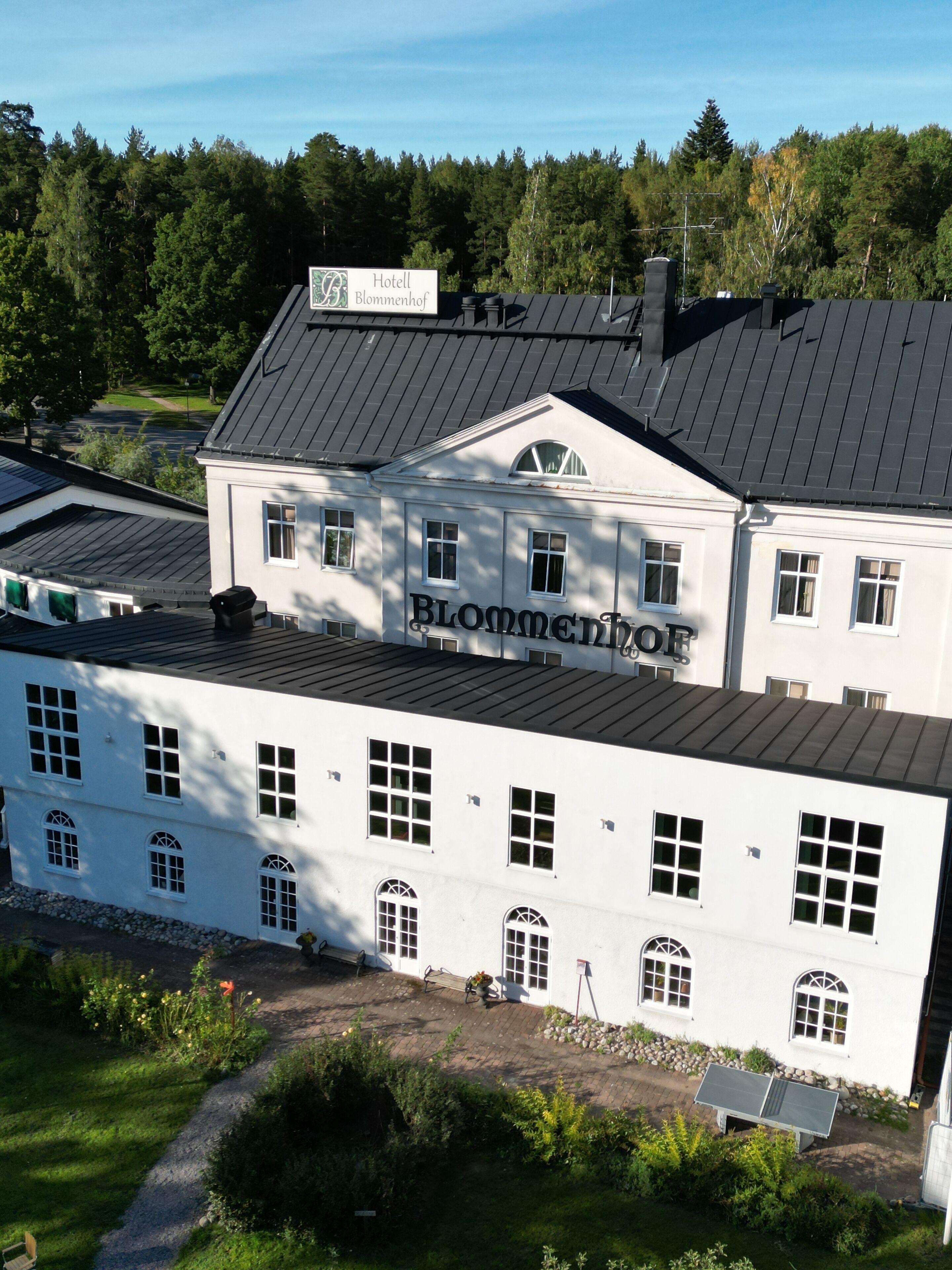 Een groot wit gebouw met de naam “Hotel Blommenhof” op de gevel, met meerdere ramen en een schuin dak.