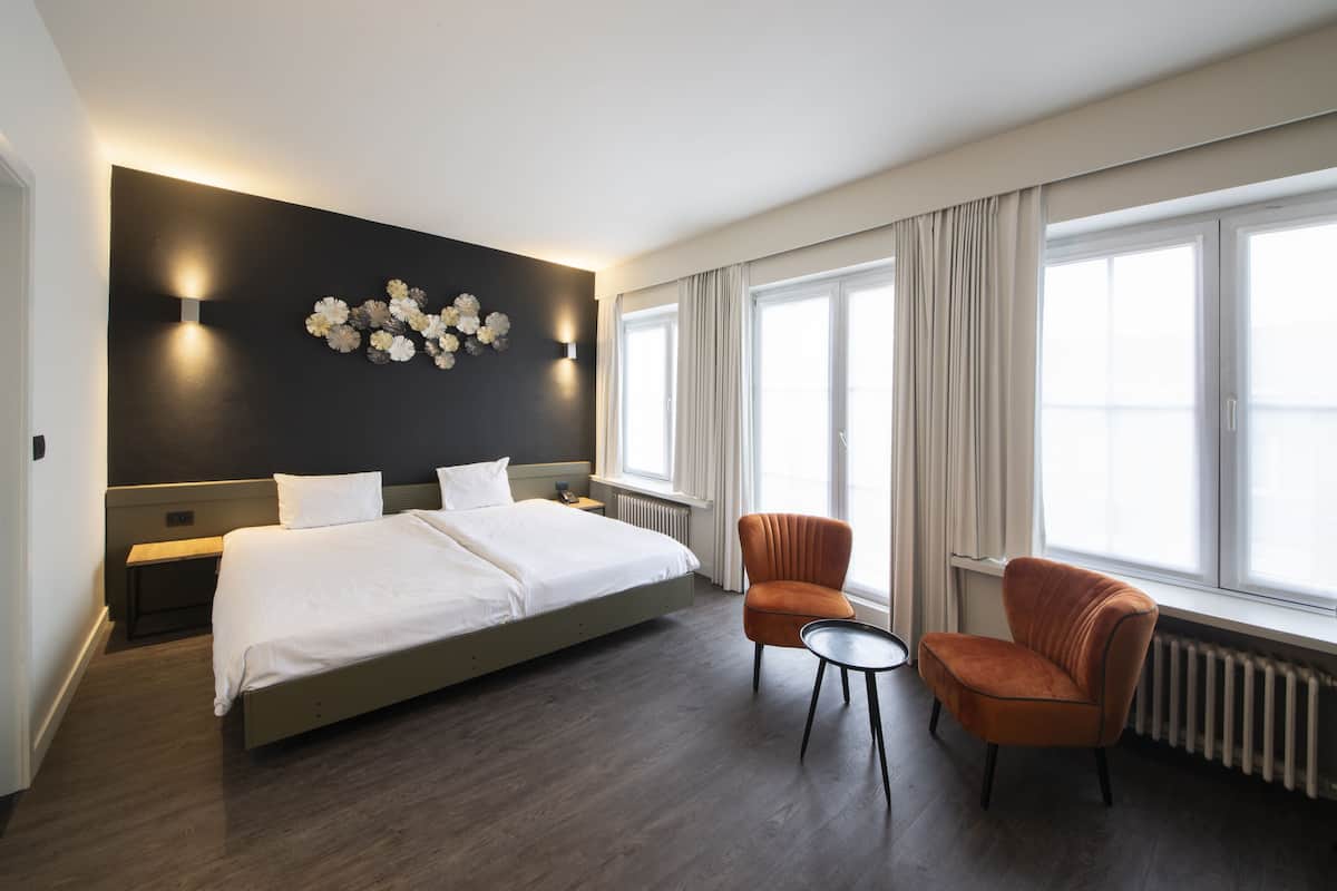A modern hotel room with a large bed, a small table, two armchairs, a wall-mounted light fixture, and a window with curtains.