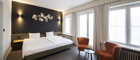 Una habitación de hotel moderna con una cama grande, una mesita pequeña, dos sillones, una lámpara de pared y un ventanal con cortinas.