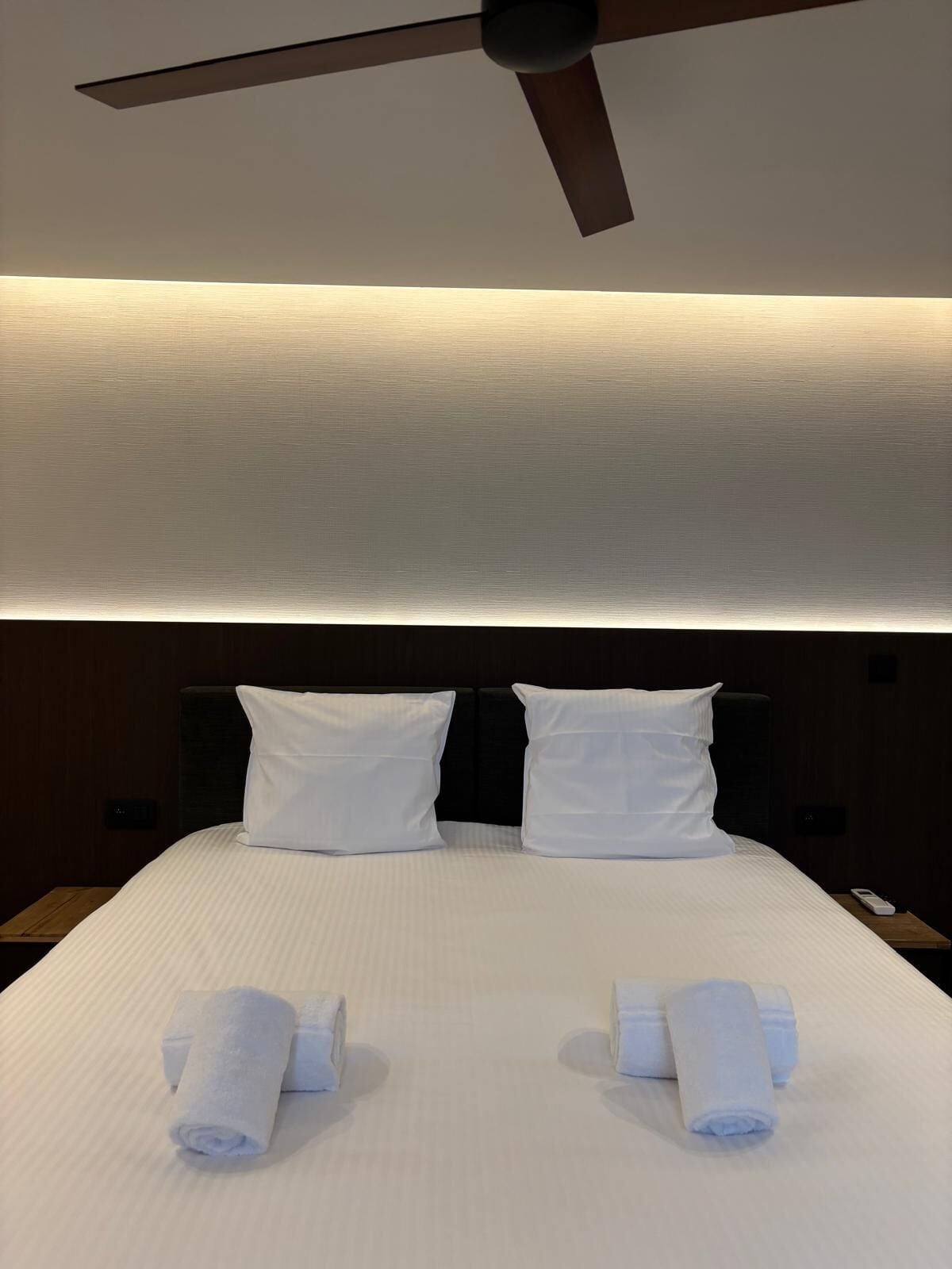 Une chambre d’hôtel avec un lit, deux oreillers, un ventilateur de plafond et deux serviettes enroulées posées sur le lit.