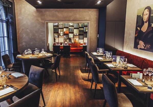 Un restaurante con mesas y sillas redondas, una zona de bar con estantes llenos de botellas y un cuadro grande en la pared.