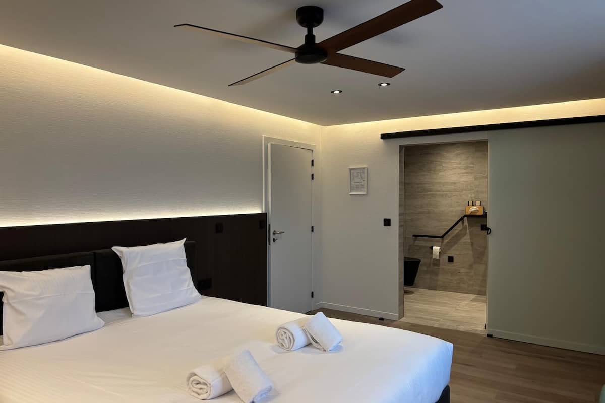 A modern hotel room with a large bed, a ceiling fan, and a view of a hallway.