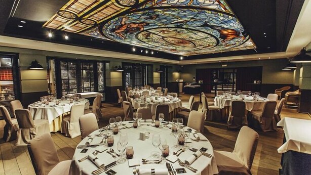 A banquet hall with round tables set for dining, a large stained glass ceiling, and a modern interior design. - Cortina Hotel (Wevelgem)