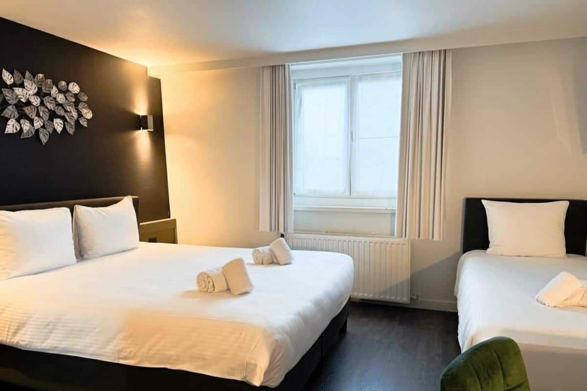 A hotel room with a large bed, a smaller bed, a window with curtains, a wall-mounted light, and a leaf-patterned wall decoration.