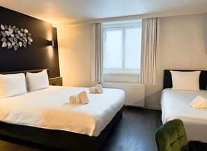 A hotel room with a large bed, a smaller bed, a window with curtains, a wall-mounted light, and a leaf-patterned wall decoration. - Cortina Hotel (Wevelgem)