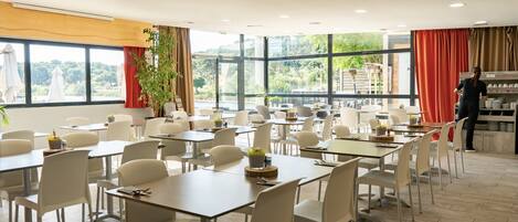 A spacious dining area with multiple tables and chairs, large windows with curtains, and a view of greenery outside.