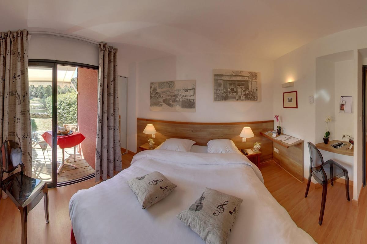 Een hotelkamer met een bed, twee nachtkastjes met lampen, een bureau met stoel, een bijzettafeltje met stoel, en een balkon met een rode stoel.
