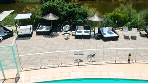 Una piscina con sillones de descanso y sombrillas, rodeada de vegetación y vista al río.
