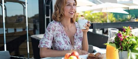 Una persona sentada en una mesa al aire libre, con un plato de comida, una bebida y un bol de fruta.